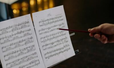 Close-up of an instructor pointing at sheet music on a stand, indoors, with blurred lights in the background.