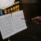 Close-up of an instructor pointing at sheet music on a stand, indoors, with blurred lights in the background.