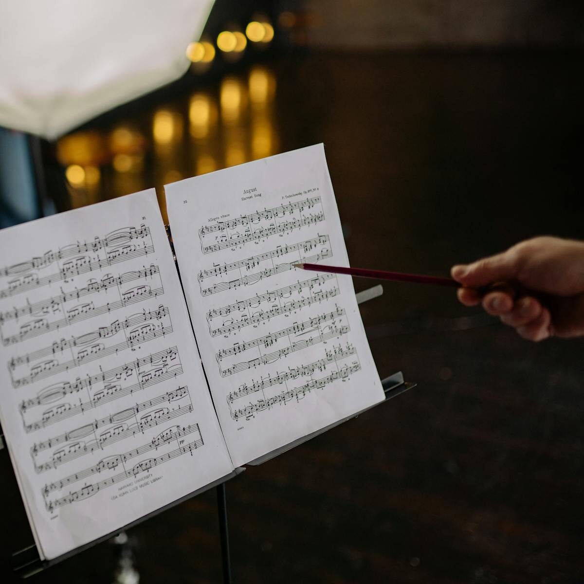 Close-up of an instructor pointing at sheet music on a stand, indoors, with blurred lights in the background.