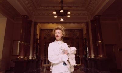 An elegant woman in a classic white dress holding a Maltese dog in a luxurious, vintage lobby.
