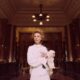 An elegant woman in a classic white dress holding a Maltese dog in a luxurious, vintage lobby.